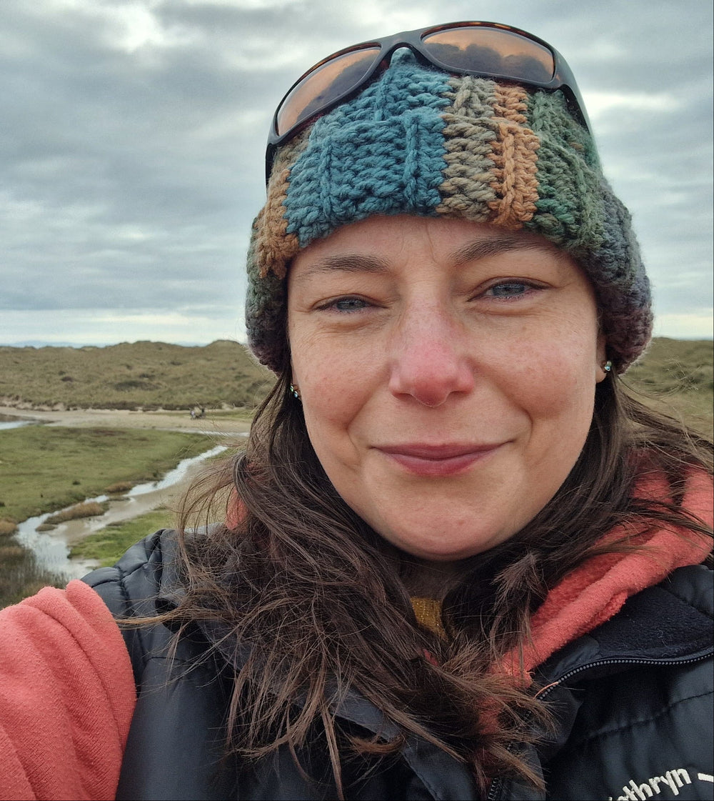 A woman taking a selfie outdoors in a coastal landscape with a winding river under a cloudy sky. She is wearing a colorful striped knit beanie, sunglasses, a dark puffer vest, and a coral fleece jacket.