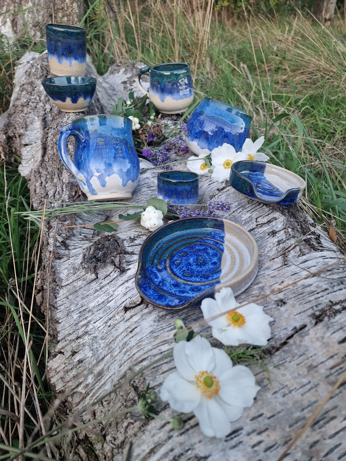 A set of handmade blue and beige glazed ceramic tableware, including mugs, cups, a pitcher, plates, and a bowl, arranged on a weathered log outdoors with white flowers and green grass in the background.
