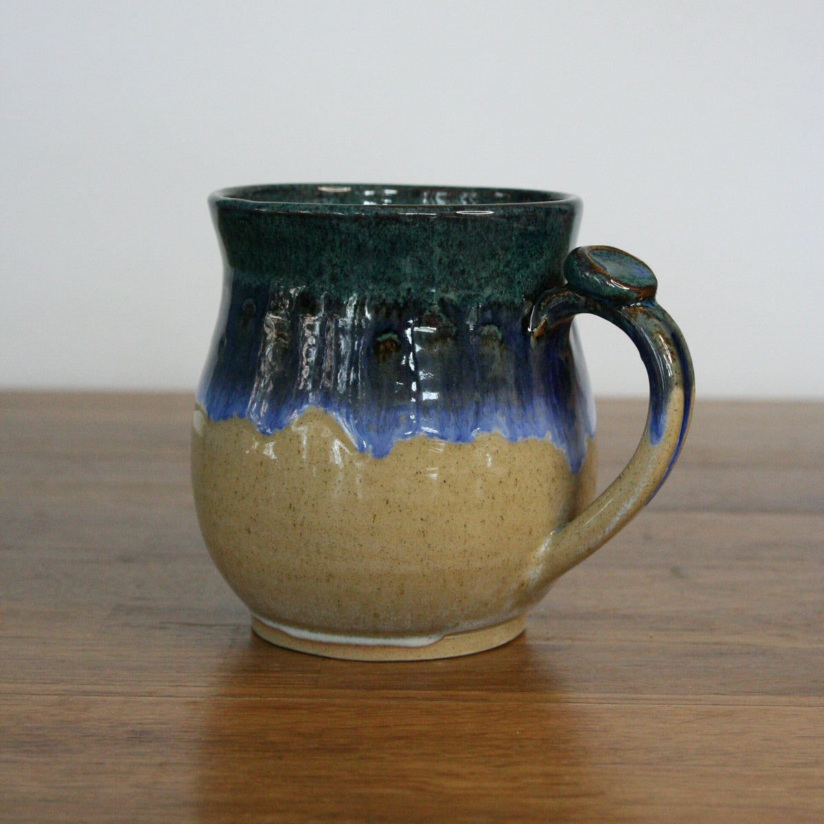 A handmade ceramic mug with a dark green and blue glaze on the upper half and a speckled beige lower half, sitting on a wooden table against a plain white wall.