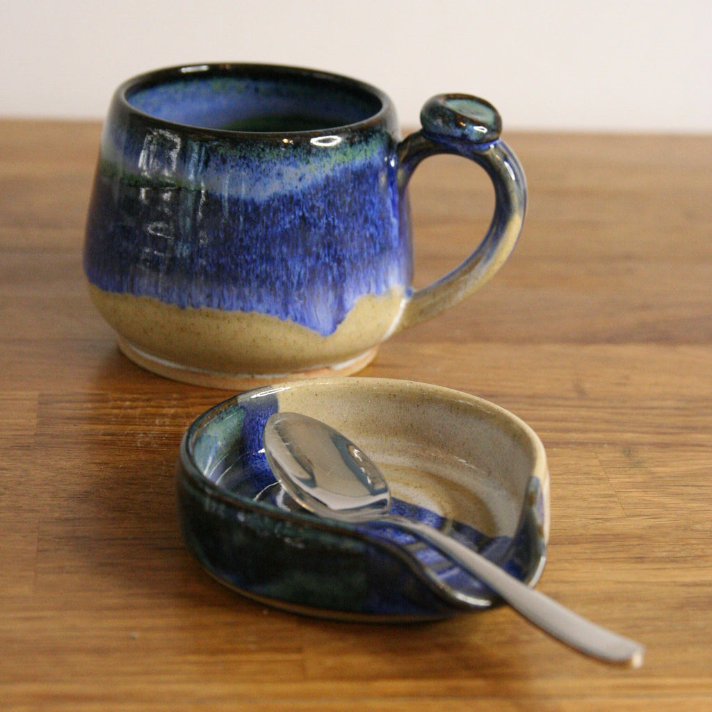 A handmade ceramic mug with a deep blue and beige glaze sits on a wooden table next to a matching ceramic spoon rest holding a silver spoon.