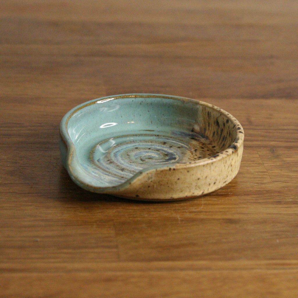 A small handmade ceramic dish with a light blue-green glazed interior and a speckled beige exterior, resting on a wooden tabletop.