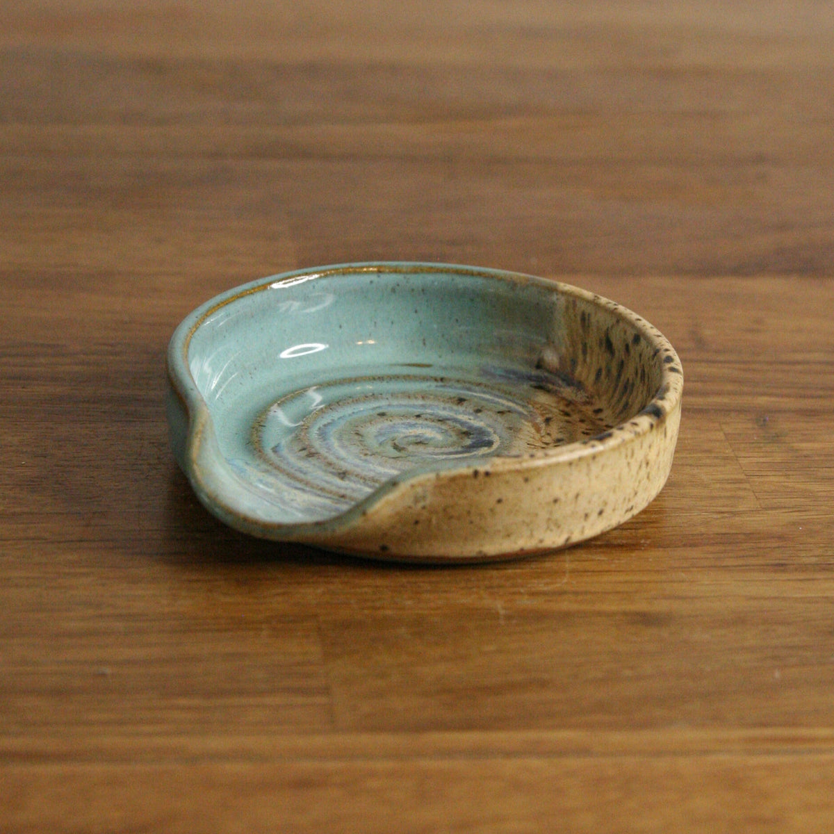 A small handmade ceramic dish with a light blue-green glazed interior and a speckled beige exterior, resting on a wooden tabletop.