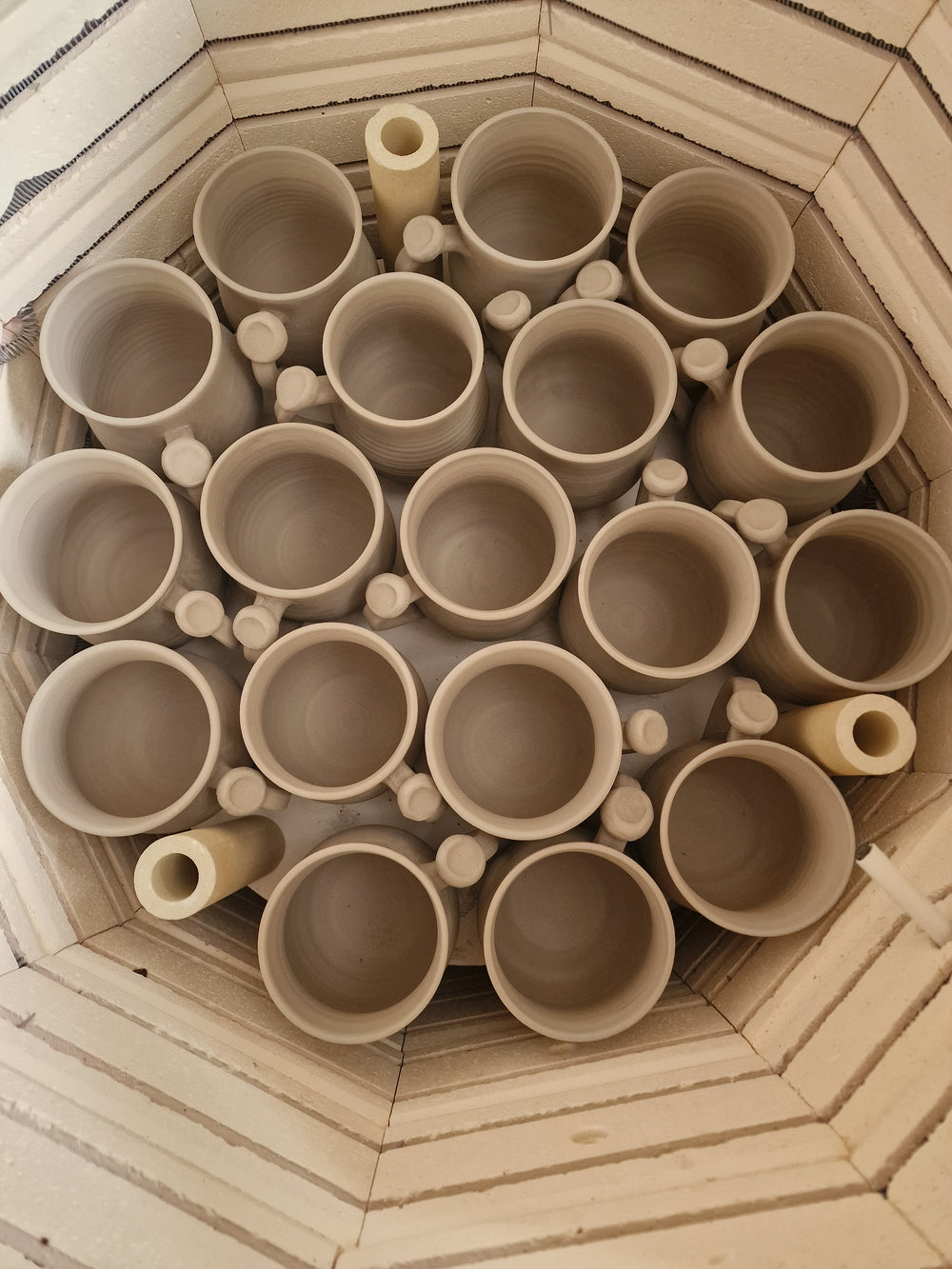 A top-down view of multiple unglazed ceramic cups arranged inside a light beige brick kiln or oven.