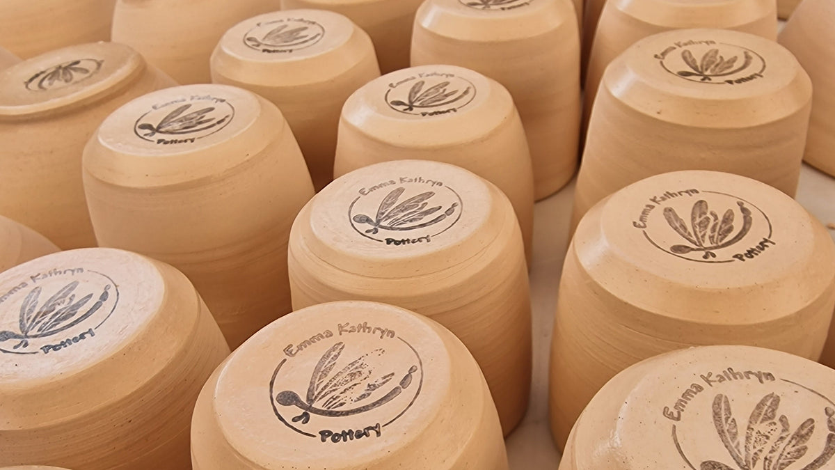 A close-up view of multiple small cylindrical ceramic pots arranged in rows, showing their light tan clay surfaces and printed circular logos on the tops. The most prominent pot in the foreground has the brand name 'Emma Kathryne Pottery' printed on it.
