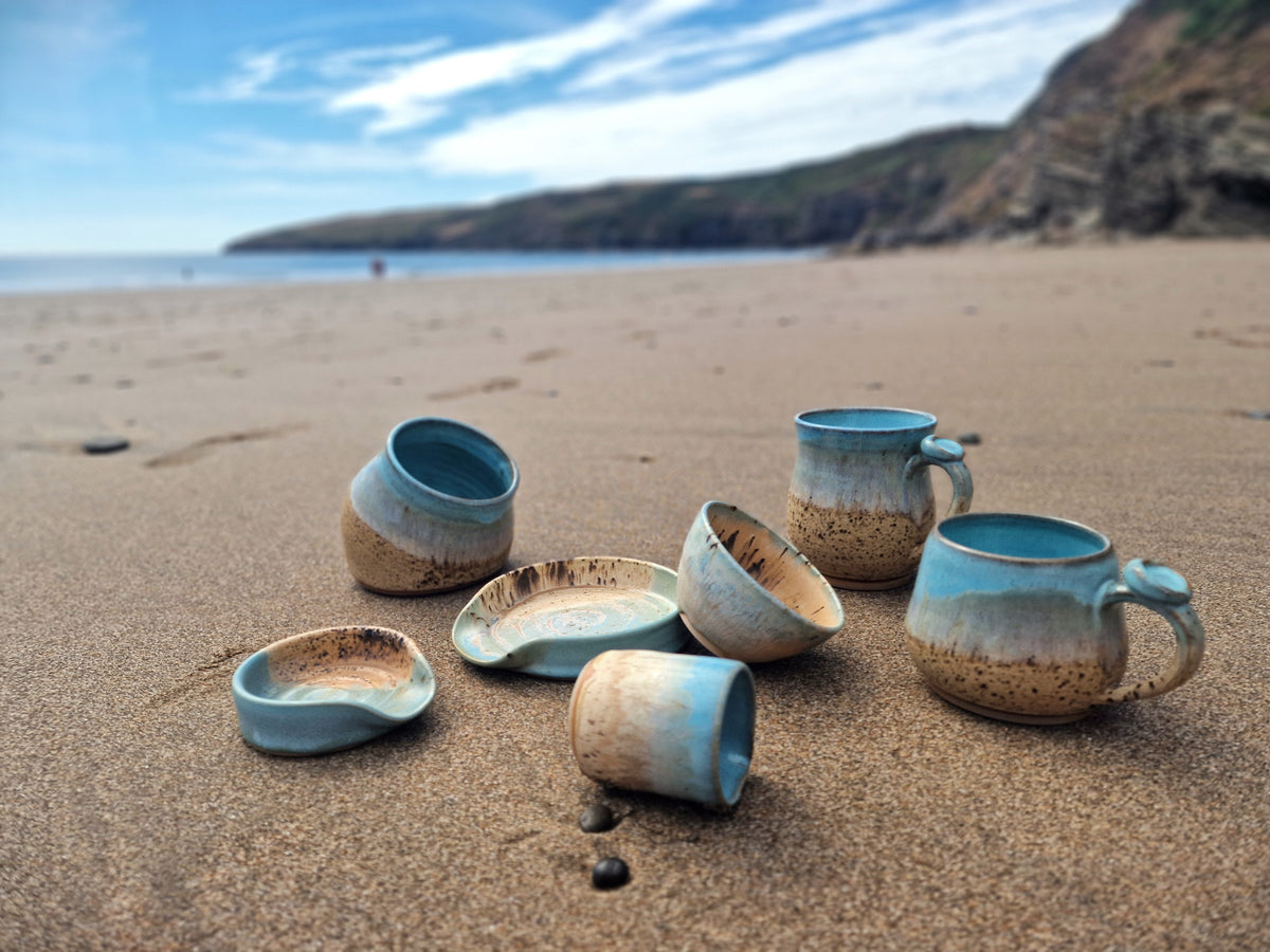 A set of handmade ceramic cups and a small plate in a coastal beach setting, featuring a blue-green glazed exterior and a speckled beige-brown interior.